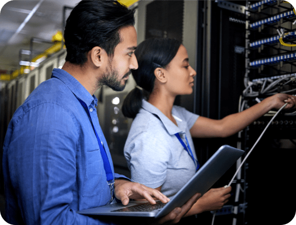Dos técnicos colaborando en un servidor dentro de un centro de datos, rodeados de filas de equipos y luces parpadeantes.