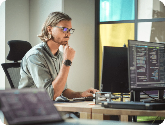 Un hombre con gafas está sentado en un escritorio, trabajando en dos computadoras una al lado de la otra.